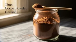 Cocoa powder in glass jar with spoon on marble surface.